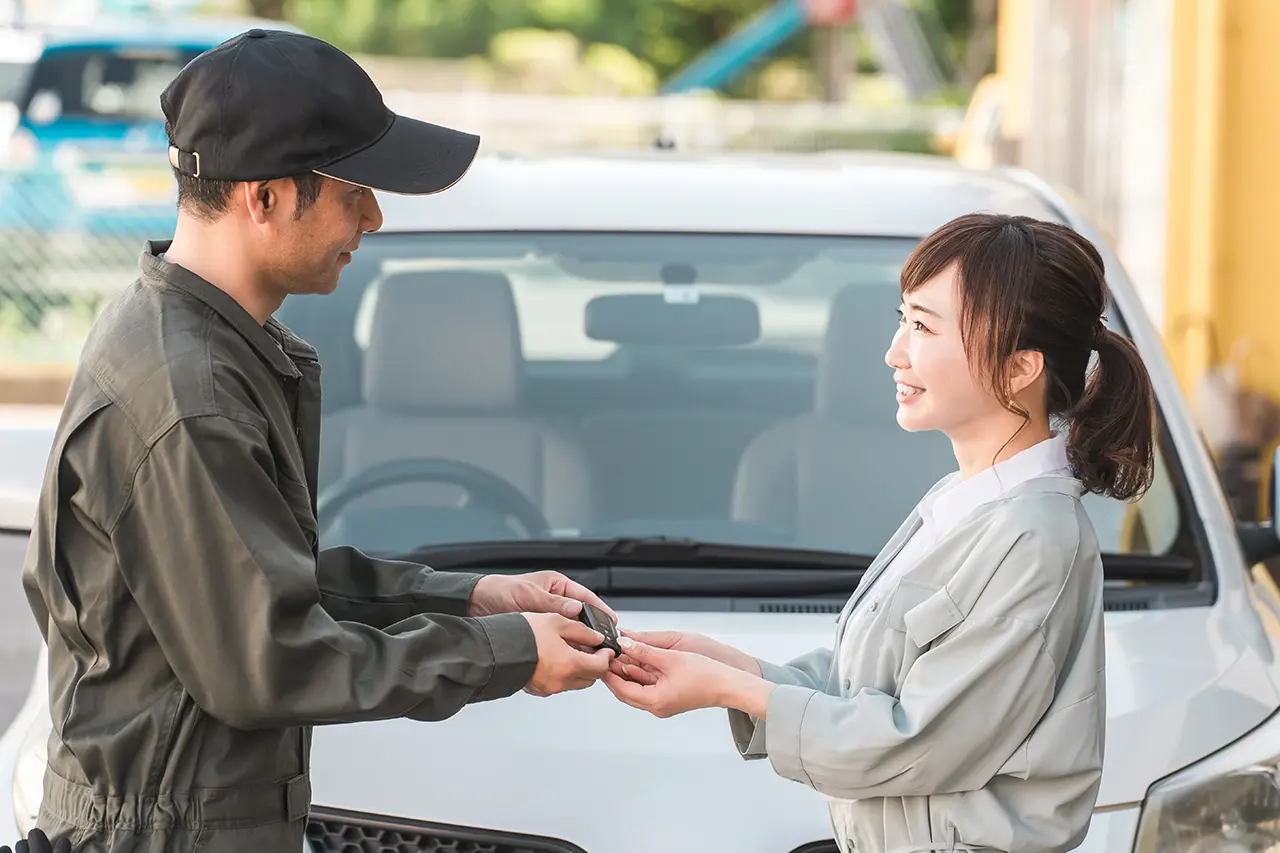 屋外で、笑顔の女性客に車の鍵を手渡す整備士